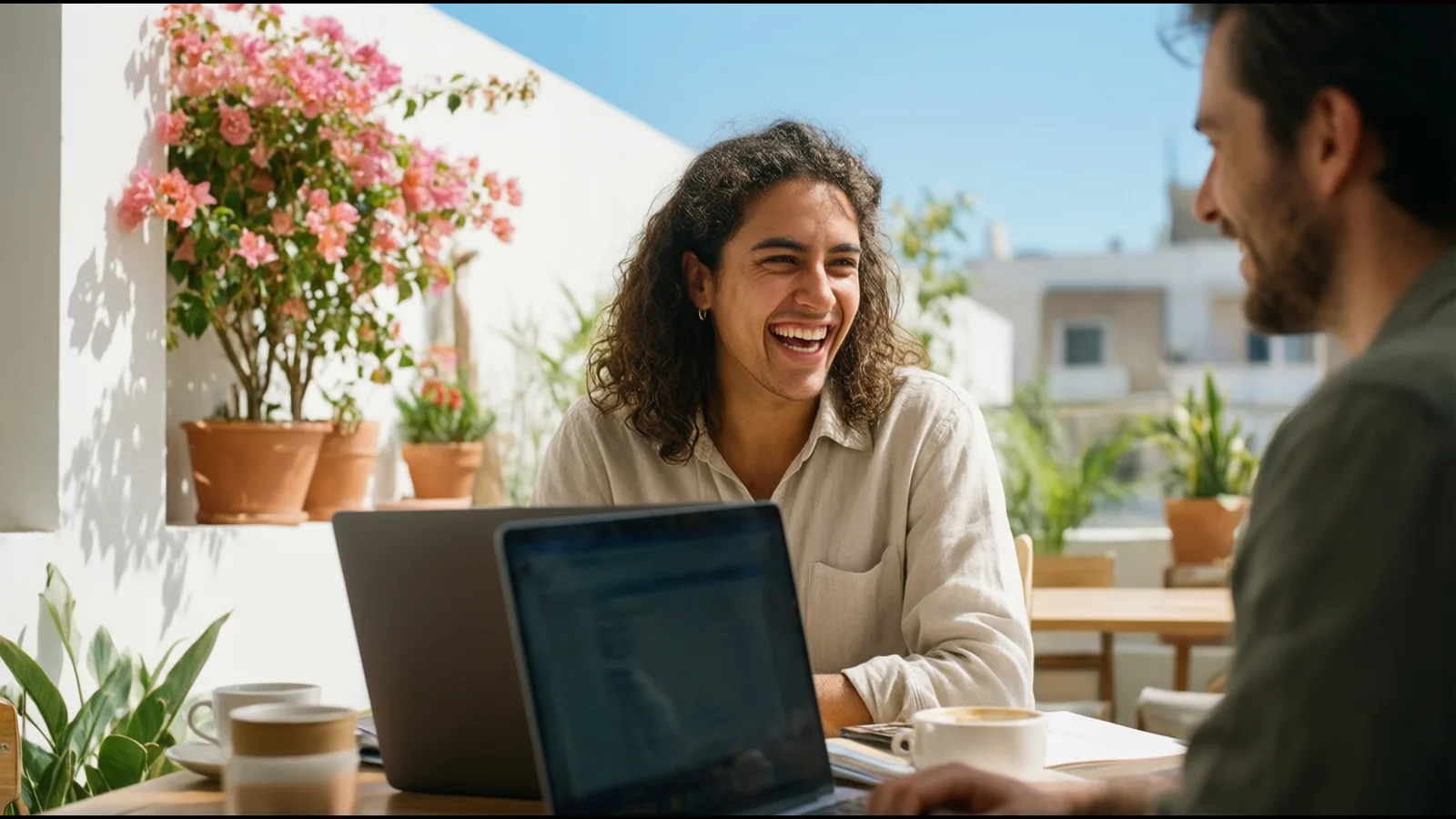 Une jeune stagiaire rit avec un collègue sur une terrasse fleurie, dans une ville du sud de l’Europe